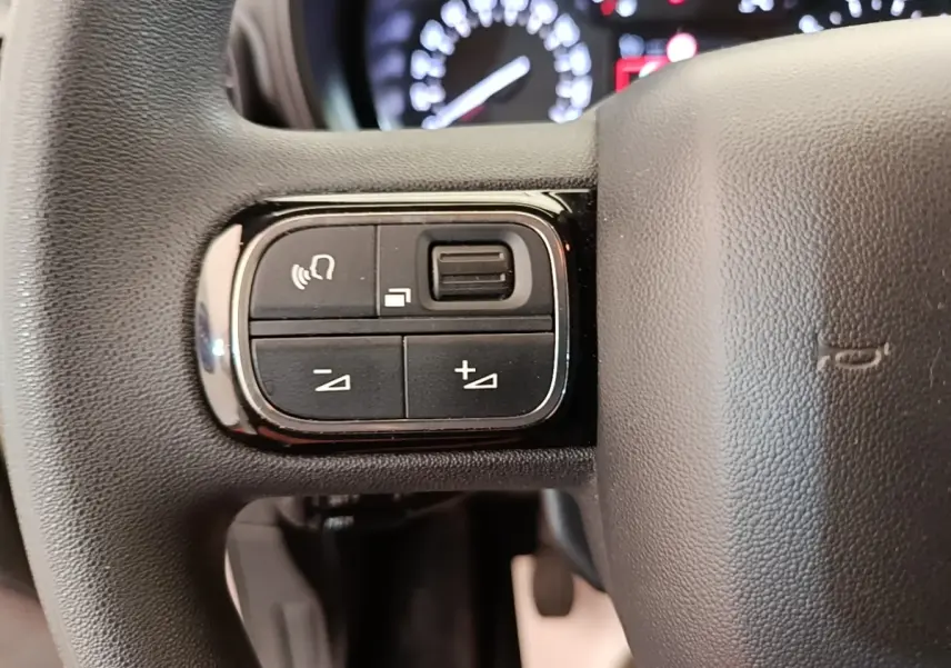 Gros plan sur les commandes multifonctions noires à gauche du volant en cuir noir du Citroën Berlingo blanc, tableau de bord flou en arrière-plan.