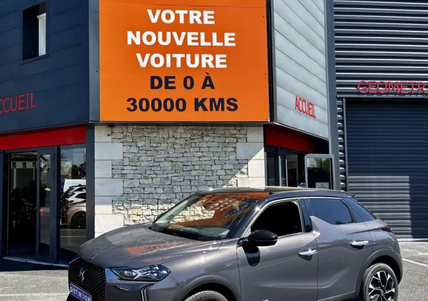 DS3 gris foncé en 3/4 avant droit, stationnée devant un bâtiment avec panneau orange Distinxion visible.
