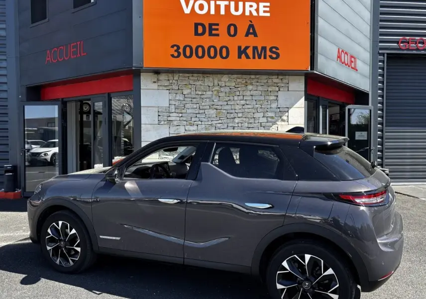 Vue de profil côté gauche d'une DS3 gris foncé garée devant un bâtiment avec panneau orange "Votre nouvelle voiture".