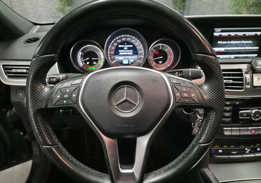 Vue rapprochée du volant en cuir noir perforé de la Mercedes Classe E E 300 BlueTEC Hybrid 2015 avec tableau de bord et écran multimédia.