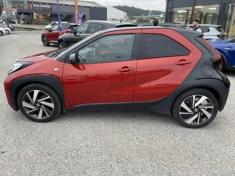Vue de profil côté gauche d'une Toyota Aygo X rouge piment avec toit noir, soulignant ses jantes distinctives et ses protections noires.