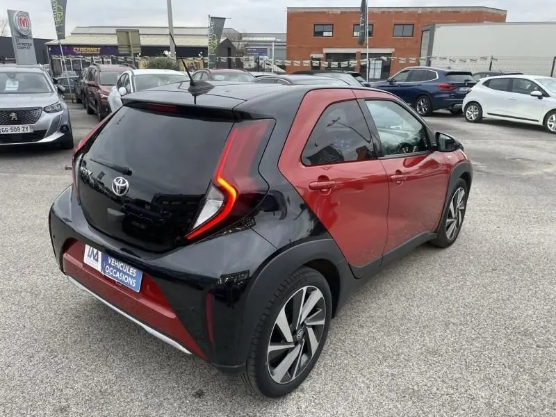 Toyota Aygo X 2023 rouge piment avec toit noir, vue 3/4 arrière droit, feux arrière en forme de boomerang allumés.