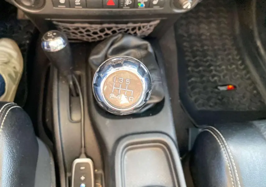 Gros plan sur le levier de vitesses manuel chromé d'une Jeep Wrangler gris clair 2012, avec console et tapis noirs visibles.