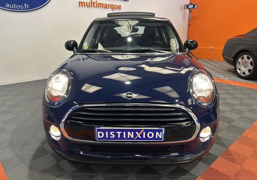 Vue avant d'une MINI 5 portes Cooper D bleu avec toit ouvrant, phares allumés, en intérieur sur sol gris et orange.