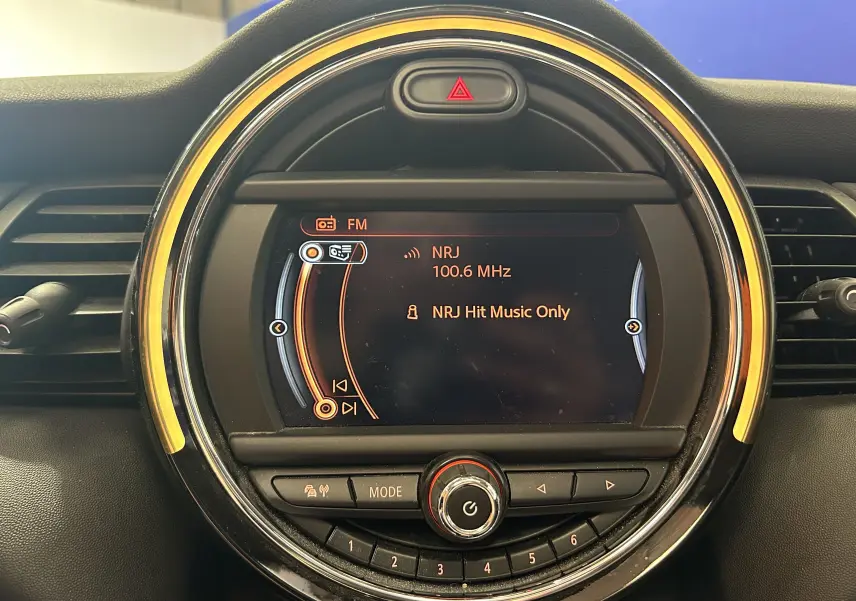 Gros plan sur l’écran central rond du tableau de bord de la MINI 5P, avec éclairage orange et affichage radio NRJ 100.6 MHz.