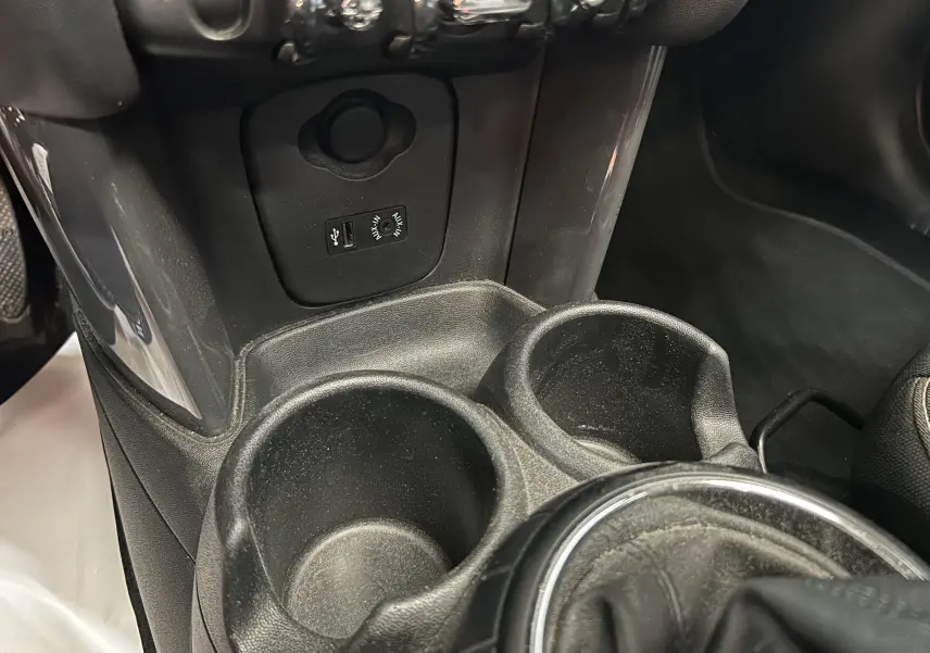 Gros plan sur la console centrale noire de la MINI 5P Cooper D 2015, montrant les porte-gobelets et prises USB/AUX.