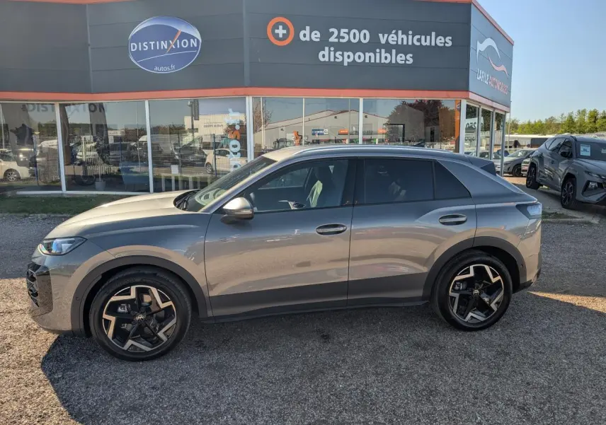 Vue de profil côté gauche d'un Volkswagen T-Roc gris métallisé avec jantes noires et toit noir, stationné devant un showroom.