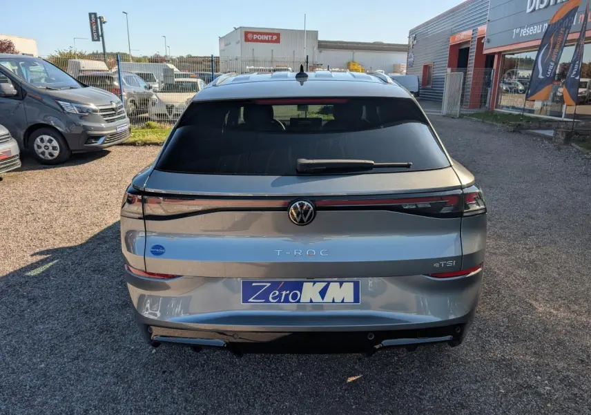 Vue arrière d'un Volkswagen T-Roc gris métallisé avec bandeau lumineux et logo VW rétroéclairé, stationné en extérieur.