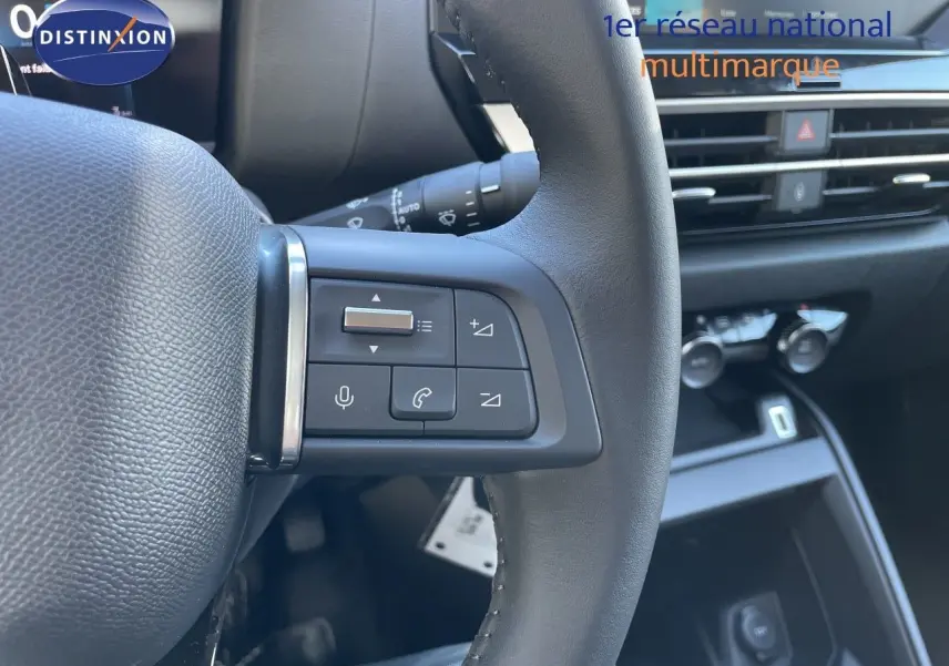 Gros plan sur les commandes gauche du volant d’une Citroën C4 hybride 2026, avec tableau de bord et console en arrière-plan.
