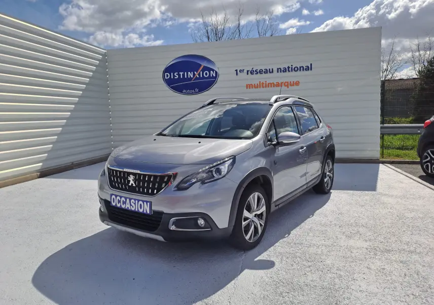 Peugeot 2008 gris en 3/4 avant droit, avec toit ouvrant et jantes alliage, stationné devant un panneau publicitaire.