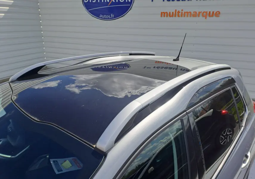 Vue en plongée du toit panoramique noir et des barres de toit argentées d'un Peugeot 2008 gris stationné près d'un mur blanc.