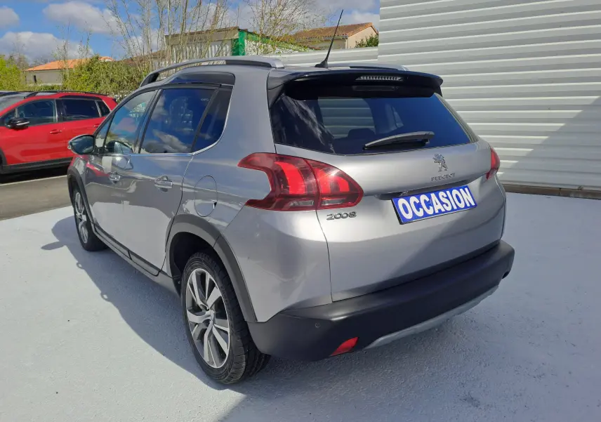 Peugeot 2008 gris vue 3/4 arrière droit, avec barres de toit et feux arrière distinctifs, sur parking extérieur.