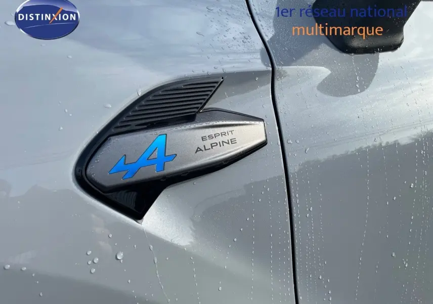 Gros plan sur l'écusson Esprit Alpine bleu sur la portière côté gauche d'une Renault Symbioz gris Rafale mouillée.