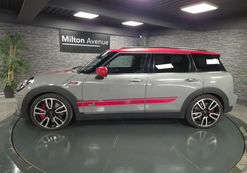 Vue de profil droit d'une MINI Clubman gris métallisé avec toit et rétroviseurs rouges, bandes décoratives sport rouges et jantes 19 pouces.