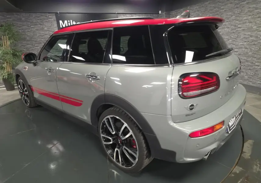 MINI Clubman John Cooper Works gris métallisé avec toit rouge, vue 3/4 arrière droite, jantes sport et bandes rouges latérales.