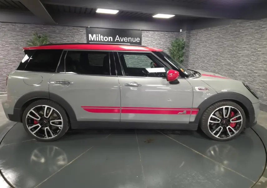 Vue de profil côté gauche d'une MINI Clubman John Cooper Works gris avec toit et détails rouges sportifs en intérieur showroom.