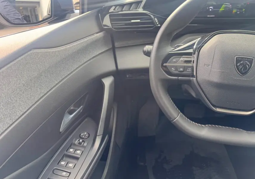 Vue intérieure côté conducteur de la Peugeot 308 gris, volant multifonction et commandes des vitres électriques visibles.
