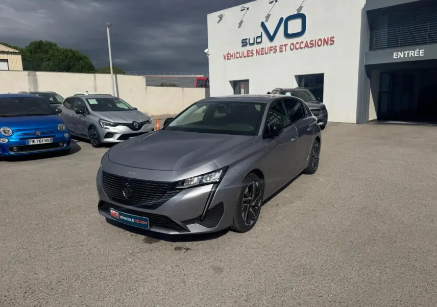 Peugeot 308 Hybrid gris vue 3/4 avant droit, jantes alliage 17 pouces, stationnée devant un bâtiment de vente auto.
