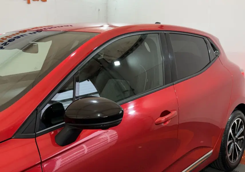 Vue latérale gauche rapprochée d’une Renault Clio rouge 2024 avec rétroviseur noir et vitres teintées.