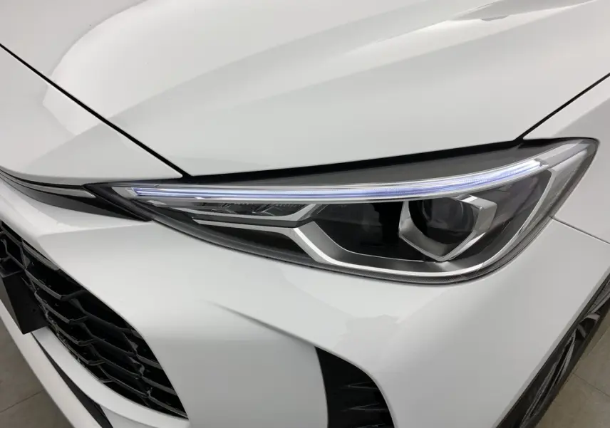 Gros plan sur le phare avant droit blanc Dover White du MG ZS 1.5 L Hybrid+ 197 ch Luxury avec calandre noire ajourée.