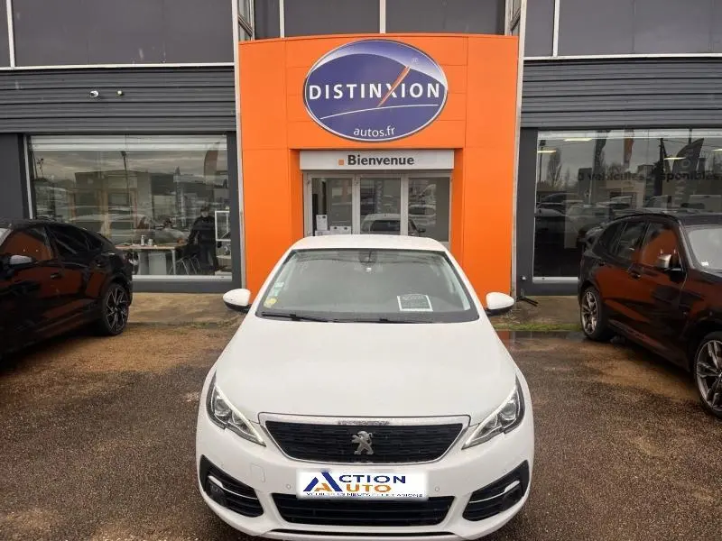 Vue avant d'une Peugeot 308 blanche Banquise garée devant une concession avec un panneau orange Distinxion.