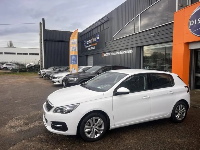 Peugeot 308 blanc banquise vue de profil droit, stationnée devant un bâtiment commercial avec d'autres voitures.