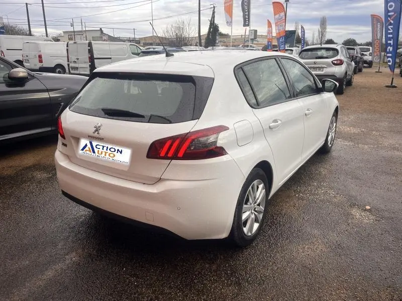 Vue 3/4 arrière droite d'une Peugeot 308 blanche Banquise avec feux arrière LED et jantes alu sur parking humide.