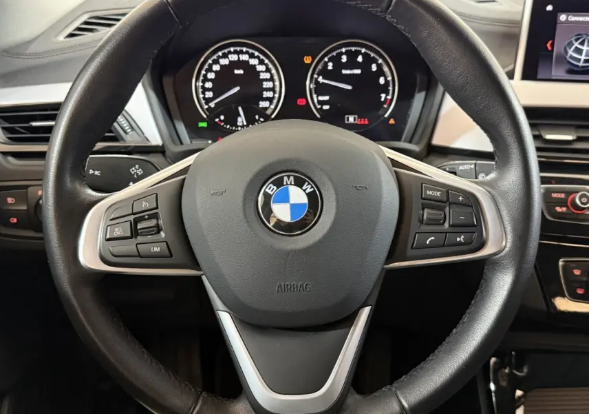 Vue rapprochée du volant cuir noir multifonctions d’une BMW X2 sDrive18i 2023 avec tableau de bord numérique en arrière-plan.