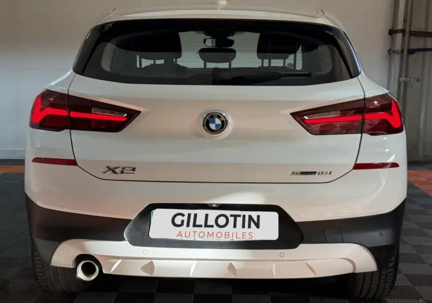 Vue arrière d’un BMW X2 sDrive18i blanc avec feux LED allumés et logo central visible dans un garage.