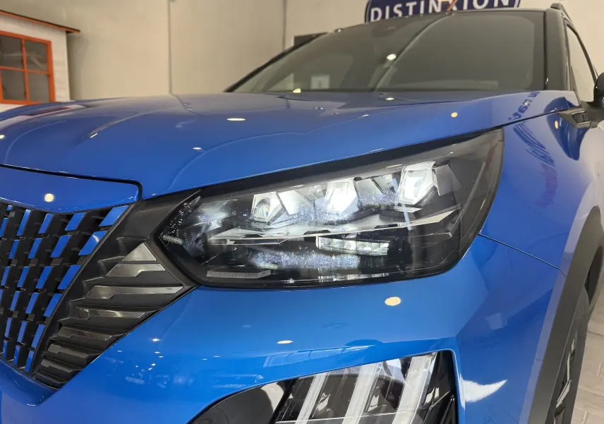 Gros plan sur le phare avant droit du Peugeot 2008 bleu Vertigo avec calandre noire et toit noir visible en arrière-plan.