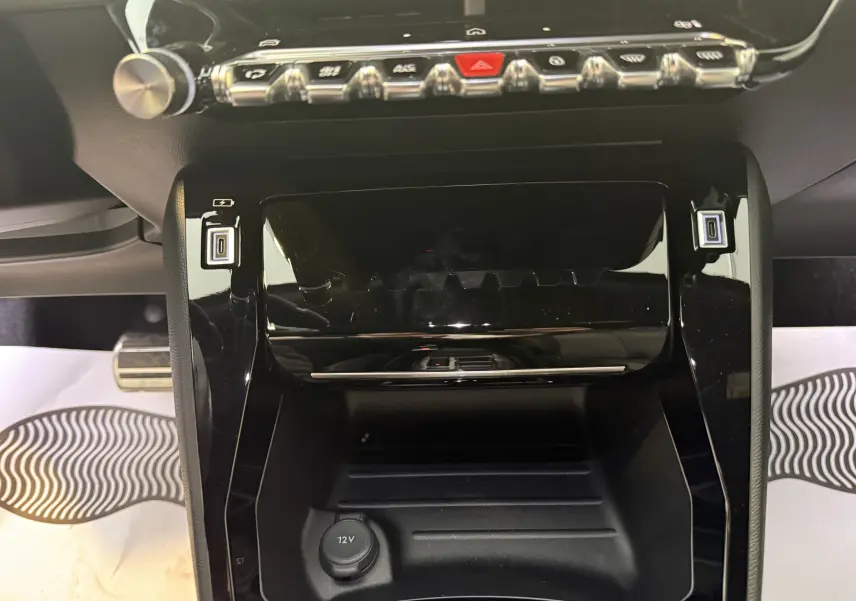 Vue rapprochée de la console centrale noire brillante du Peugeot 2008 1.2 PureTech 130ch GT avec boutons de commande et prise 12V.