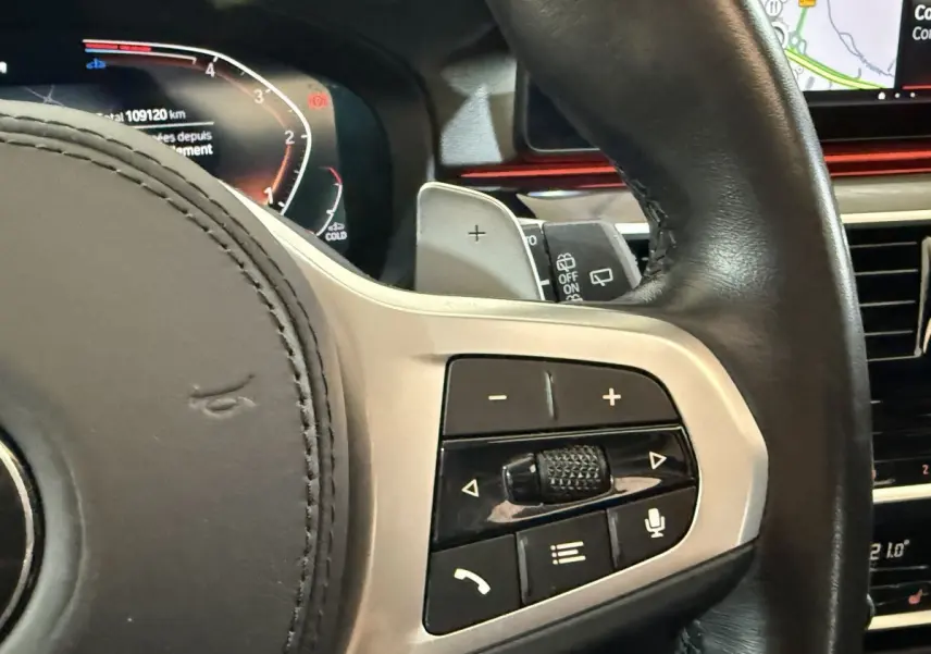 Gros plan sur le volant cuir noir de la BMW Série 5 Touring 520d, avec commandes multifonctions et écran digital en arrière-plan.