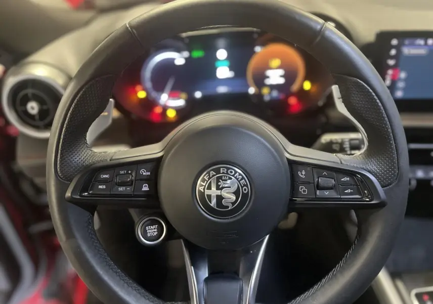 Volant sport en cuir noir avec commandes intégrées et bouton start/stop, tableau de bord digital lumineux en arrière-plan.