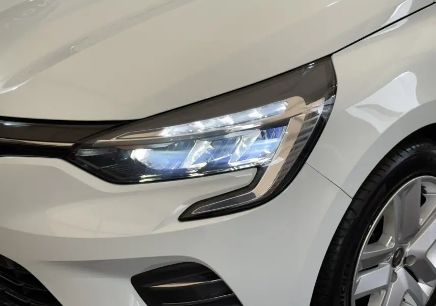 Gros plan sur le phare avant droit allumé d’une Renault Clio Business blanche avec jante alliage visible.