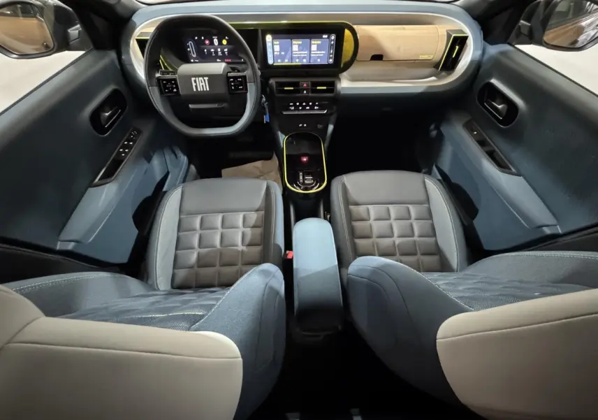 Vue intérieure avant de la Fiat Grande Panda 2025, montrant tableau de bord marron clair et sièges bi-ton gris et beige.