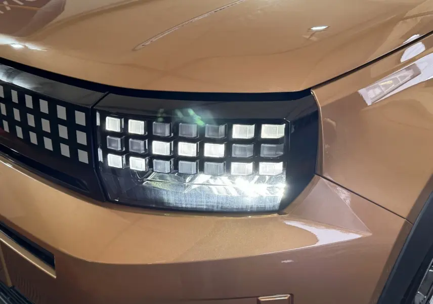 Gros plan sur le phare avant droit allumé de la FIAT Grande Panda marron avec calandre à motifs carrés.