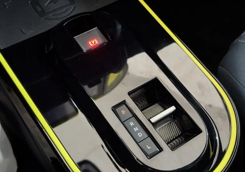 Gros plan sur la console centrale noire brillante avec bouton frein électrique rouge et sélecteur de boîte auto entouré de jaune.