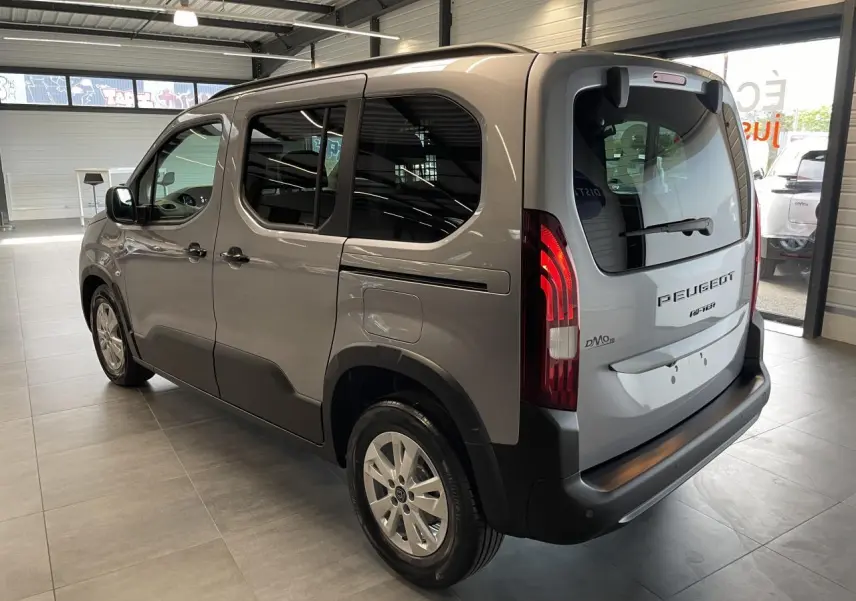 Peugeot Rifter gris Artense vu en 3/4 arrière droit, avec vitres teintées et feux arrière allumés en intérieur showroom.