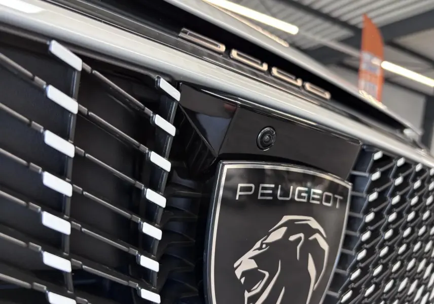 Gros plan sur la calandre noire laquée avec logo Peugeot argenté et caméra frontale du Peugeot 3008 blanc.