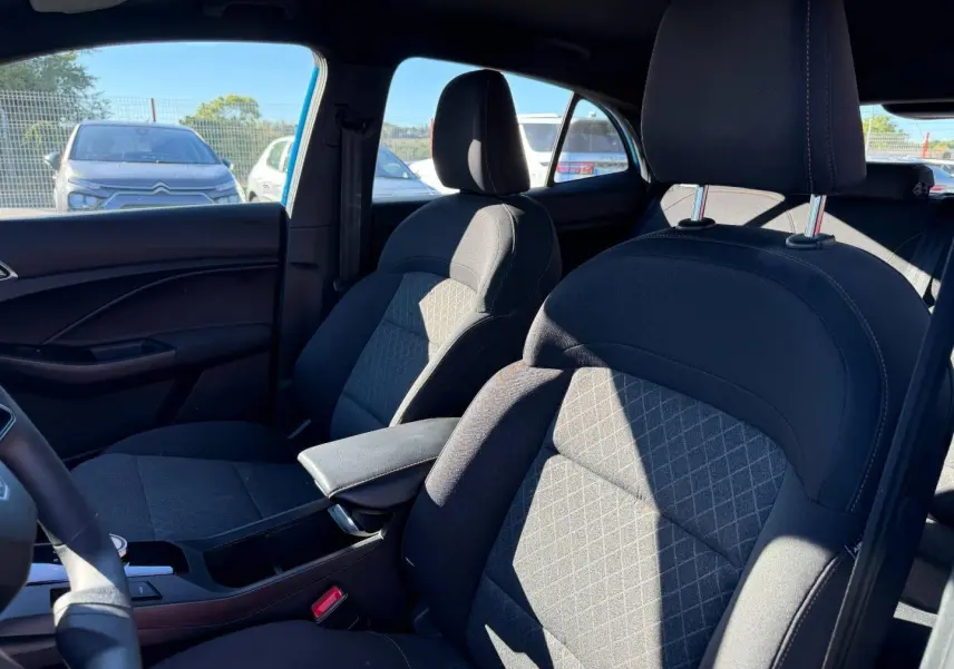 Vue intérieure côté avant droit du MG3 1.5 Hybrid+ 2024 avec sièges tissu noir et accoudoir central.