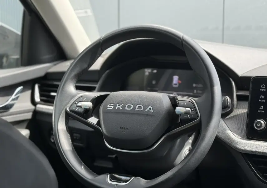 Vue rapprochée du volant cuir noir multifonctions avec logo SKODA, tableau de bord et écran tactile visibles en arrière-plan.