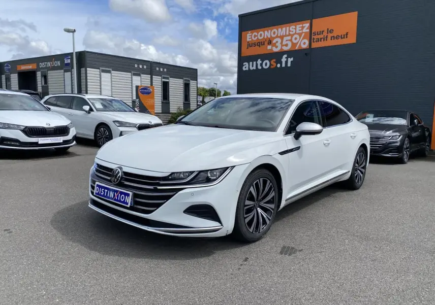 Volkswagen Arteon blanc pur vu en 3/4 avant droit sur parking devant un bâtiment commercial.