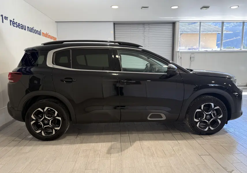 Profil latéral droit d'un Citroën C5 Aircross gris Artense métal avec jantes alliage et barres de toit visibles en intérieur showroom.
