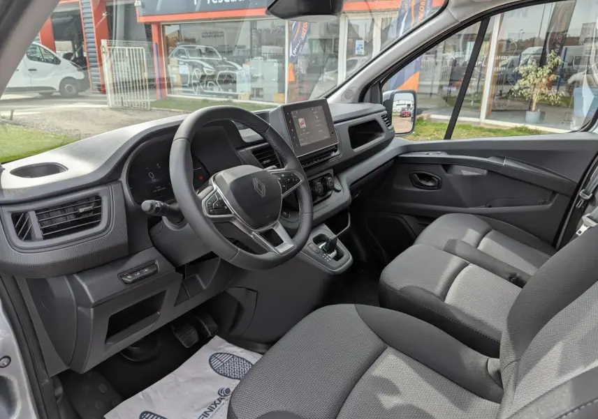 Vue intérieure côté conducteur du Renault Trafic Fourgon gris, montrant le volant, la console centrale et la banquette passager 2 places.