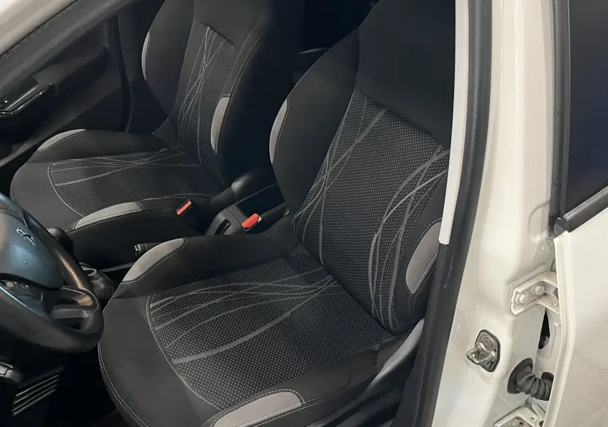 Vue intérieure côté conducteur d'une Peugeot 208 blanche de 2014, sièges tissu noir avec motifs gris et volant visible.