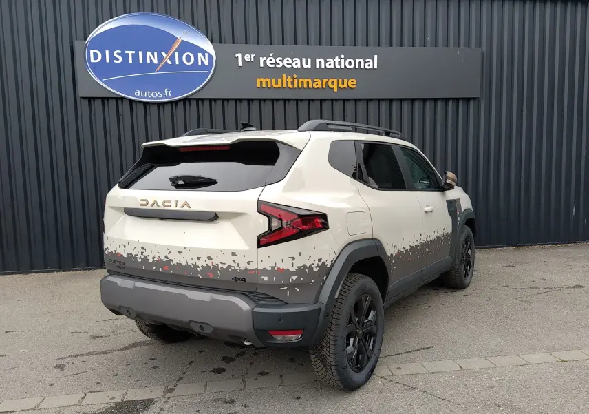 Vue 3/4 arrière droite du Dacia Duster 2026 hybride bicarburation en couleur Sandstone avec motifs pixelisés gris et rouge.