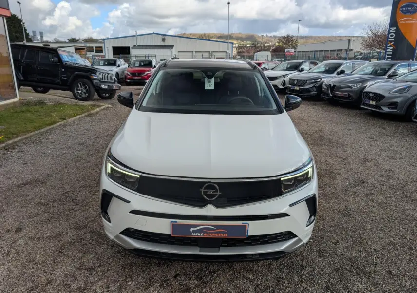 Vue avant d'un Opel Grandland blanc métallisé avec calandre noire et phares LED allumés sur un parking extérieur.