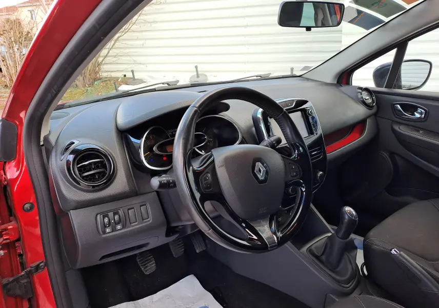 Vue intérieure côté conducteur de la Renault Clio IV rouge, montrant le volant, tableau de bord et levier de vitesses manuel.