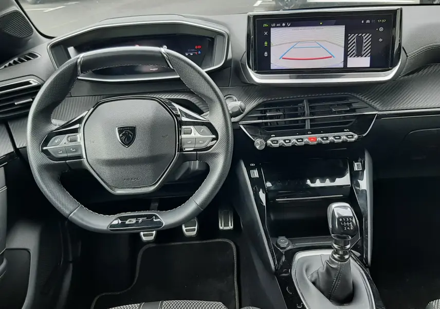 Vue intérieure avant de la Peugeot 208 GT 1.2 essence 2025, volant GT, tableau de bord numérique et écran tactile avec caméra de recul.