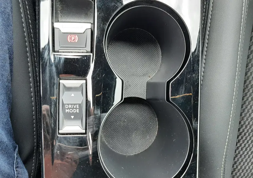 Gros plan sur la console centrale noire brillante de la Peugeot 208 1.2 100CH, avec porte-gobelets et boutons de frein à main et mode de conduite.